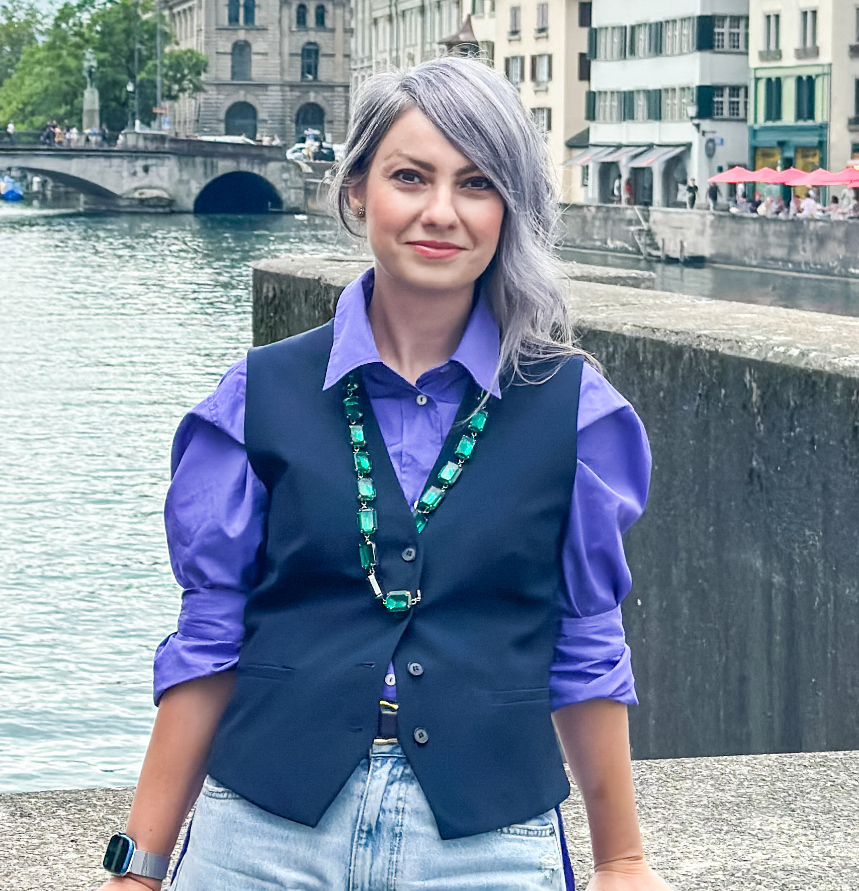 Tünde Macarie in a navy vest and purple shirt beside a river, with a stone bridge and old buildings behind her