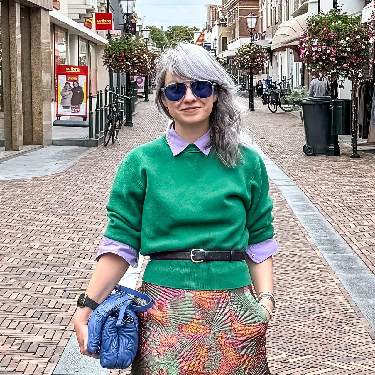 Tünde Macarie in a green sweater and blue sunglasses on a brick shopping street with hanging flower baskets