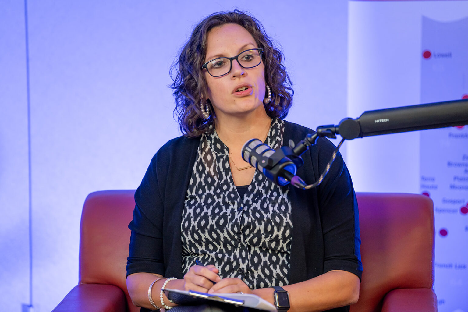 Close-up of a panelist with curly hair and dark-framed glasses speaking into a microphone while holding a clipboard at the NALHE Indiana Chapter Café con Leche podcast panel